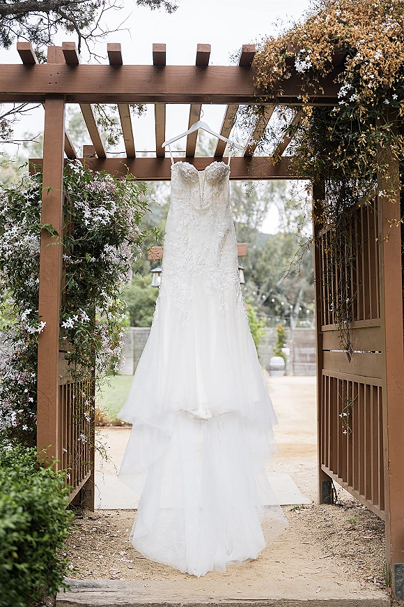 Wedding dress hanging on a hanger, strapless lace bodice with tiered tulle skirt under a wood pergola with garden greenery behind