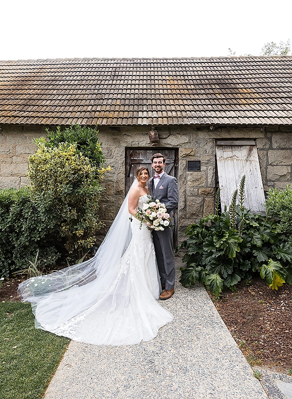 Couple portrait of bride in lace dress with long veil holding bouquet beside groom in gray suit by a stone building and rustic wooden door