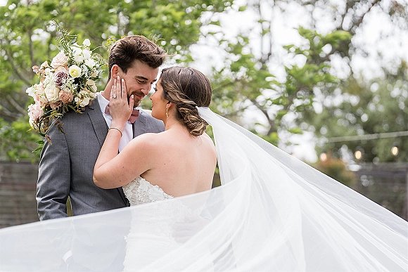 Couple portrait of bride touching groom’s face as wedding veil blows, her lace dress and bouquet beside a stone wall with string lights
