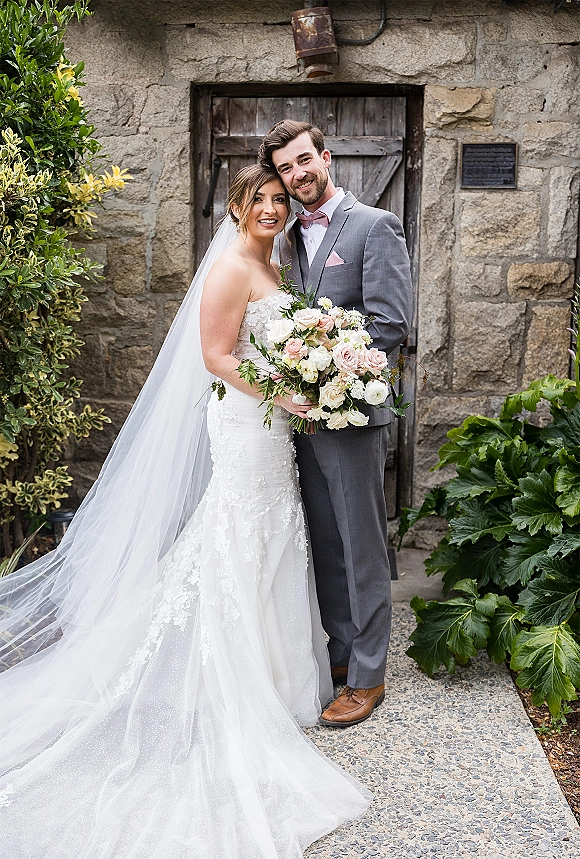 Couple portrait of bride and groom portrait hugging by a stone wall and rustic wooden door, bride holding bouquet with a long veil