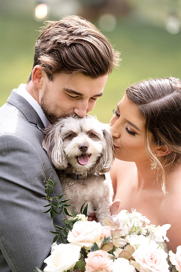 Wedding couple with dog sharing kisses while holding a small white pup and blush rose bouquet, framed by softly blurred outdoor greenery
