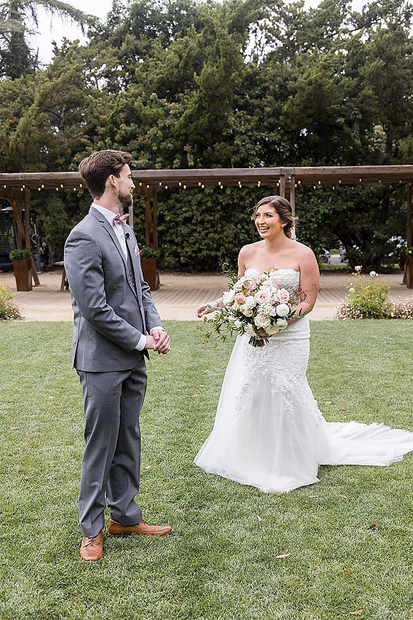 First look moment as groom in gray suit reacts to bride in strapless lace dress holding a rose bouquet under pergola string lights