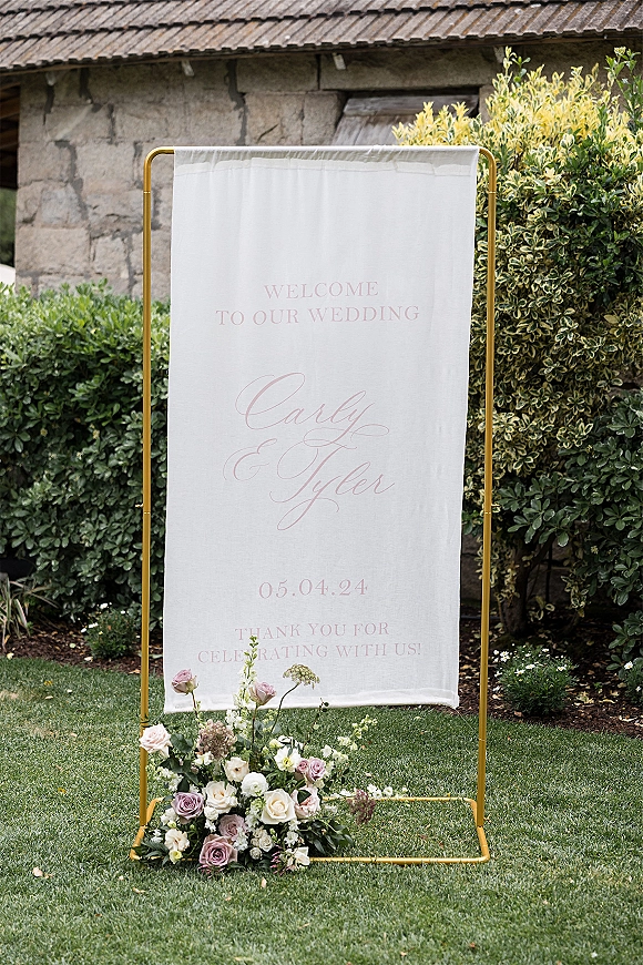 Wedding welcome sign on a gold metal stand with a fabric banner and rose floral arrangement, set on a lawn by garden shrubs and stone wall