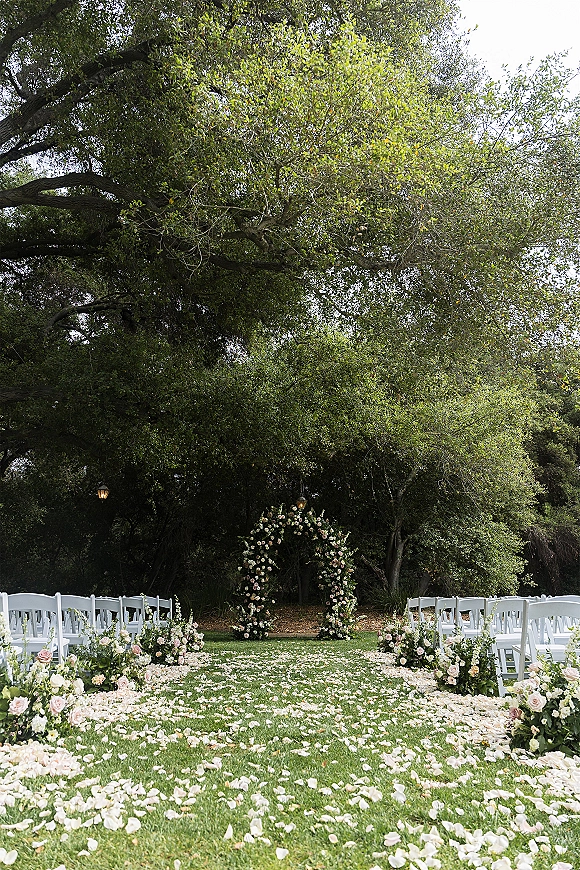 Ceremony setup for an outdoor wedding ceremony with a circular floral arch, rose petal aisle, lanterns, and white chairs on a lawn under trees