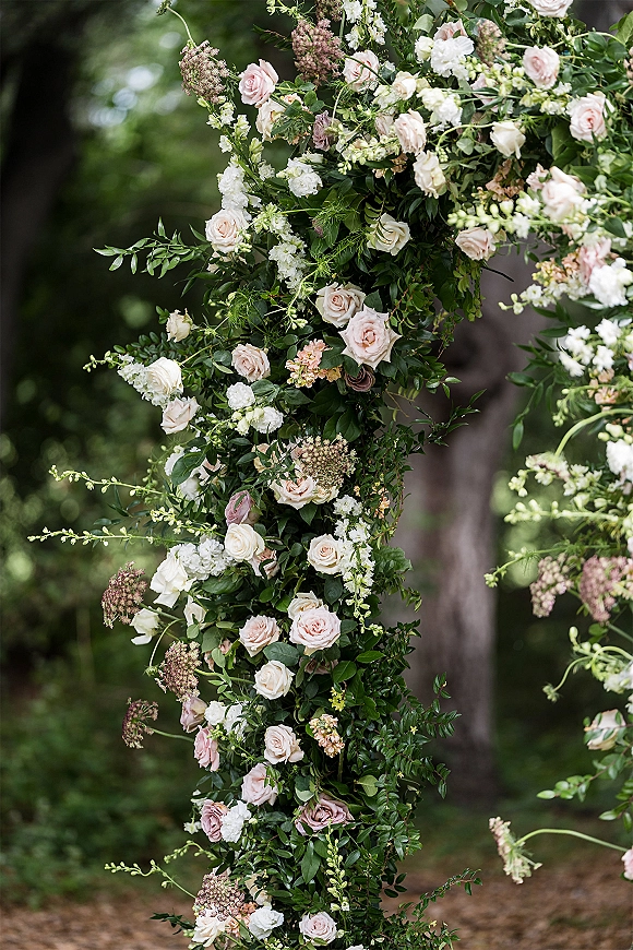 Wedding floral arch with roses and greenery, blush and white blooms in an asymmetrical design set against garden trees outdoors