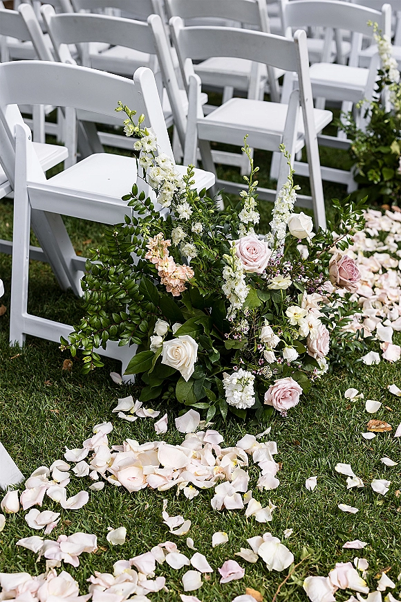 Ceremony aisle decor with wedding aisle flowers in a low ground floral arrangement beside white folding chairs on a grass lawn