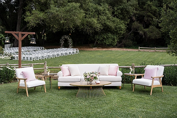 Wedding lounge setup with outdoor wedding lounge seating, white sofa and pastel pillows around a gold coffee table on a grassy lawn