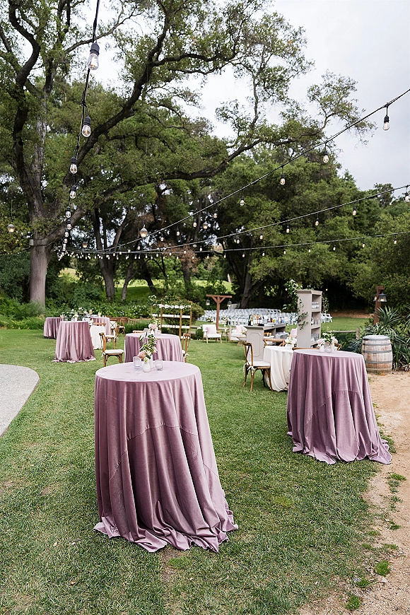 Outdoor wedding reception with string lights overhead, round cocktail tables in mauve linens, floral centerpieces, and a lawn setting