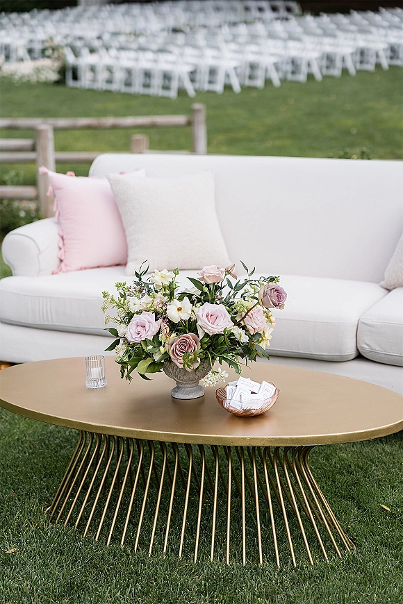 Wedding lounge seating with a white outdoor sofa and blush pillow around a gold coffee table, set on a lawn near ceremony chairs