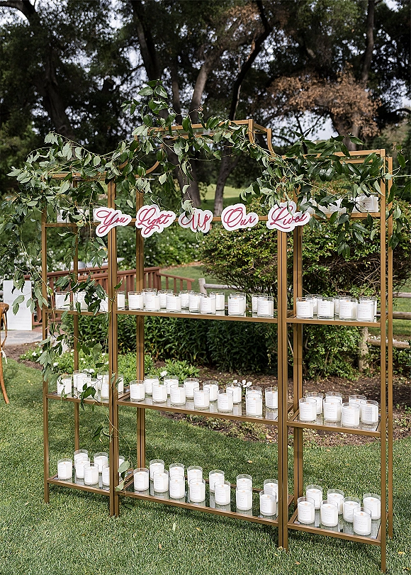 Wedding candle display on gold metal shelving with glass jar candles and greenery garland, topped by a hanging sign on a lawn backdrop