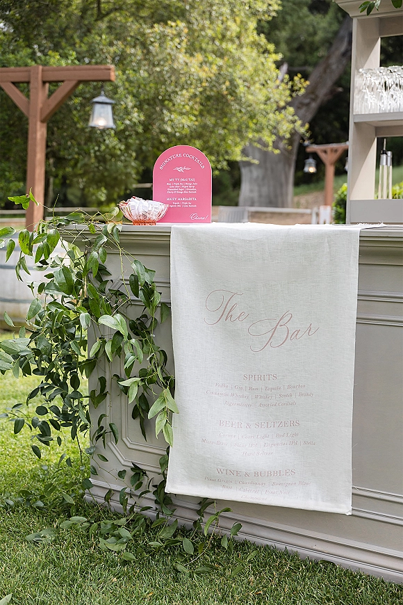 Wedding bar setup with a wedding bar menu sign and greenery garland on the counter, hanging lanterns and glassware shelf in a garden setting