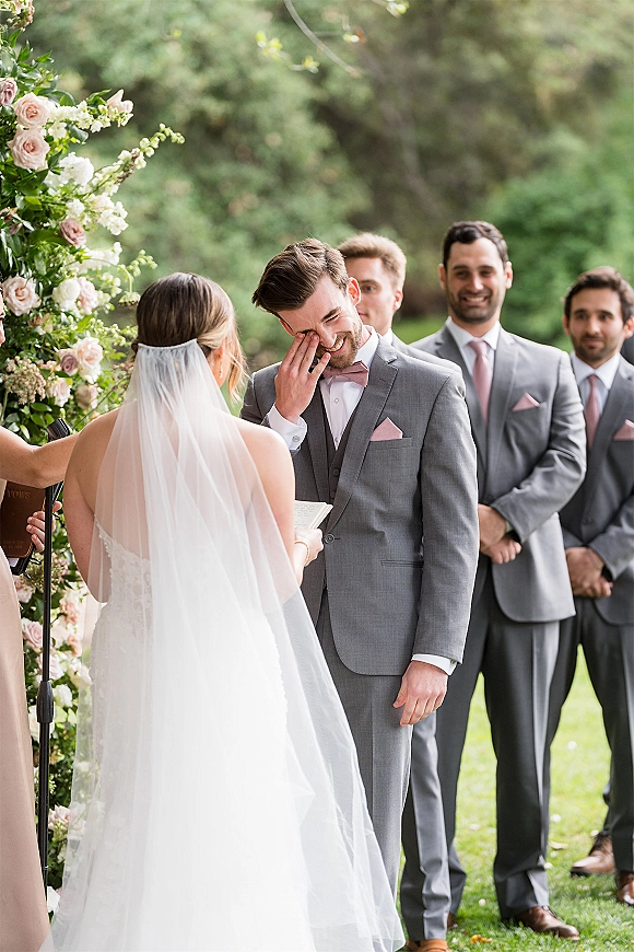 Wedding vows as bride reading vows faces groom in grey suit under rose and greenery floral arch on a garden lawn, veil trailing behind