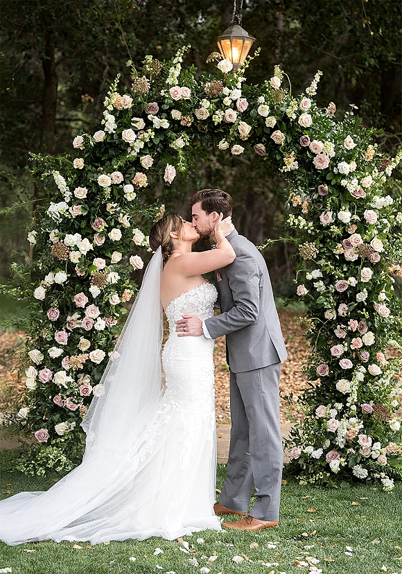 Wedding kiss portrait of bride and groom kissing beneath a round rose-and-greenery floral arch with a hanging lantern on a garden lawn