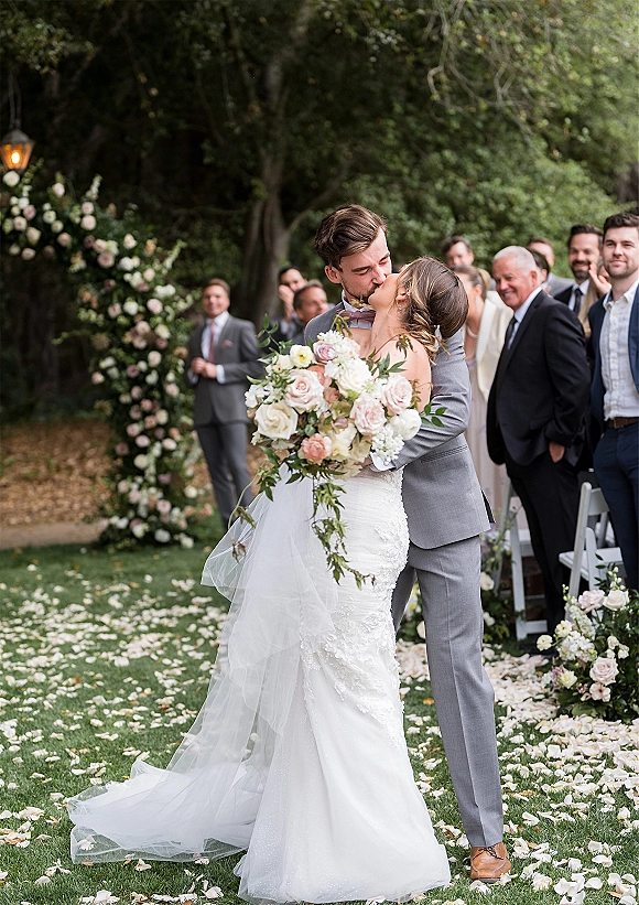Wedding kiss as bride in lace dress and veil holds bouquet, kissing groom in gray suit beneath floral arch on garden aisle with guests