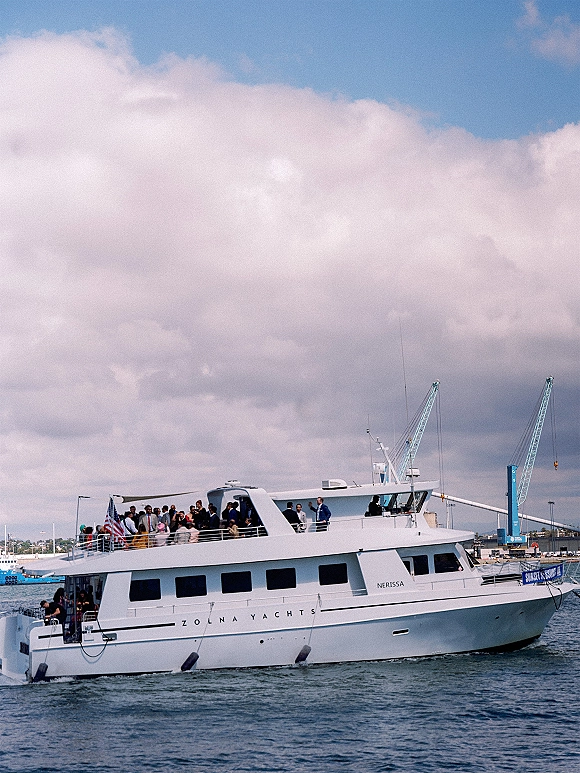 Wedding yacht docked in a harbor, white vessel with upper deck railing and American flag under cloudy sky with port cranes nearby