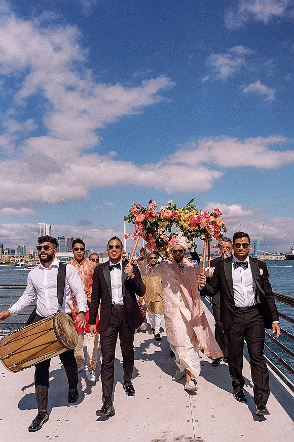 Baraat procession at an Indian wedding baraat with men in turbans and tuxedos under a floral canopy on a waterfront pier, city skyline behind