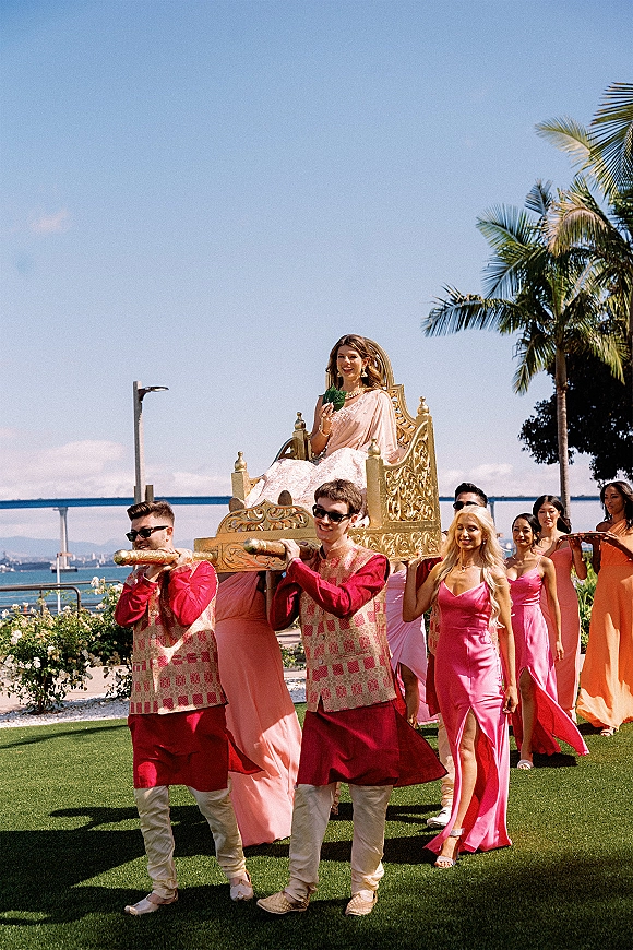 Wedding processional with bridal entrance chair as bridesmaids in pink dresses and men in embroidered vests walk on a lawn by palm trees and ocean view