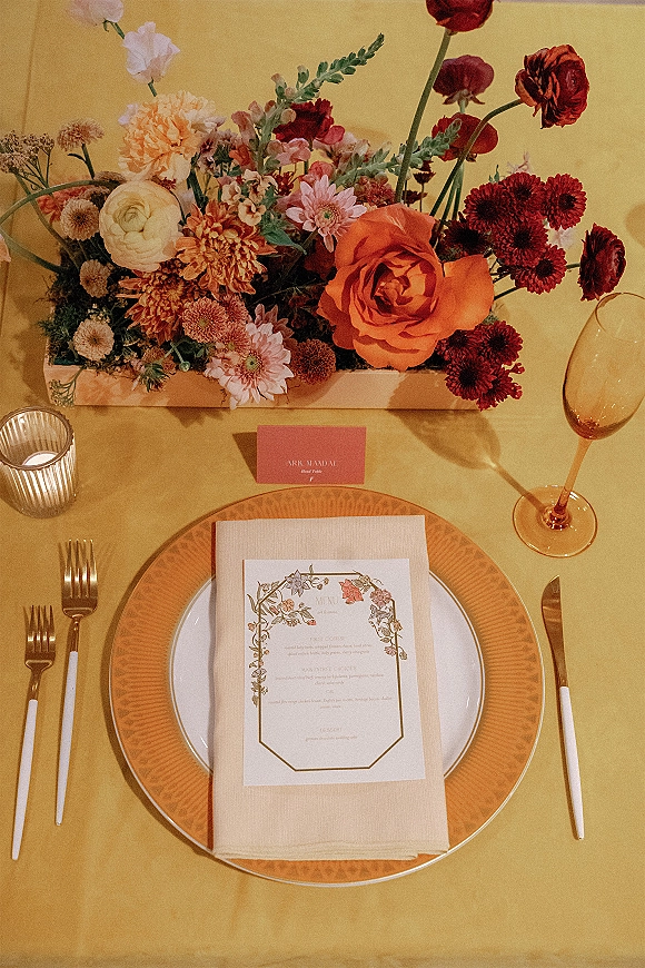Reception tablescape with wedding place setting, gold flatware, layered plates, menu and place cards, roses centerpiece on yellow tablecloth