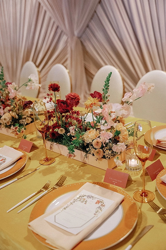 Reception tablescape with wedding table centerpiece, gold chargers, champagne flutes, and a votive candle before a draped fabric backdrop