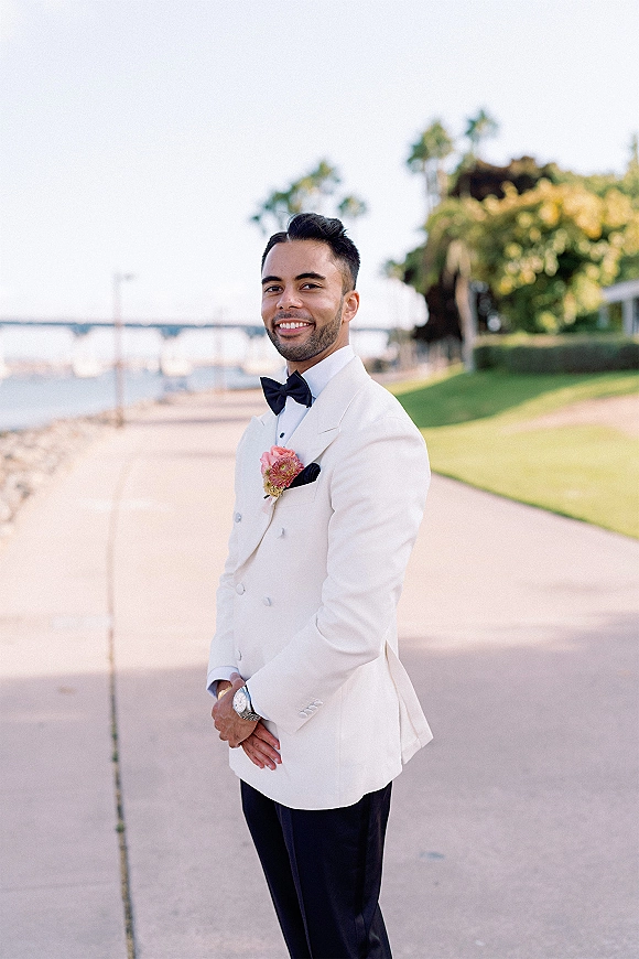 Groom portrait in a white tuxedo jacket with black bow tie, boutonniere, and wristwatch, standing on a waterfront walkway by a bridge