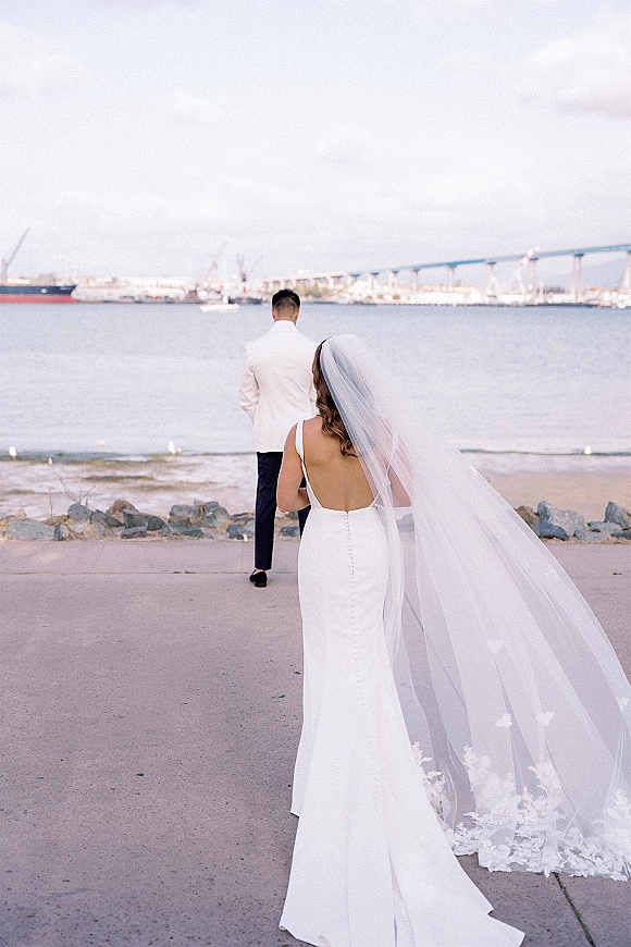 Wedding first look as bride in a fitted dress approaches groom in a white dinner jacket on a waterfront walkway with bridge under cloudy sky
