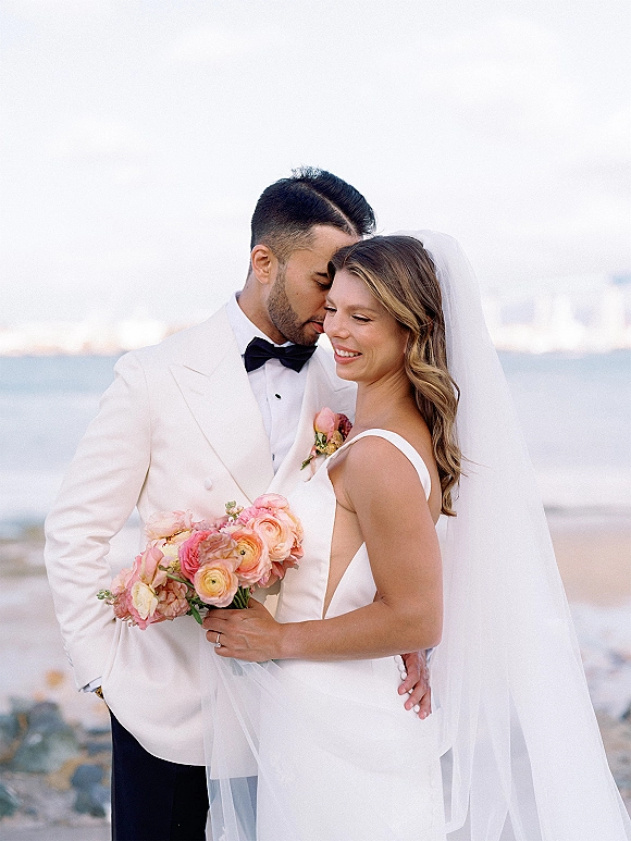 Couple portrait of bride and groom embrace as he whispers, her veil and bouquet flowing, on a beach with ocean and rocks behind