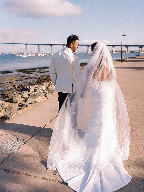 Couple portrait of bride and groom walking away, her lace-edged veil and gown train flowing on a waterfront promenade by sailboats