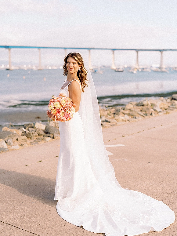 Bridal portrait of a bride holding a bouquet with a long veil and sleek wedding dress, standing on a rocky waterfront shore near a bridge