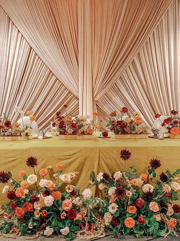 Reception sweetheart table with a sweetheart table backdrop, yellow linen, votive candles, table lamps, and meadow-style florals on the floor