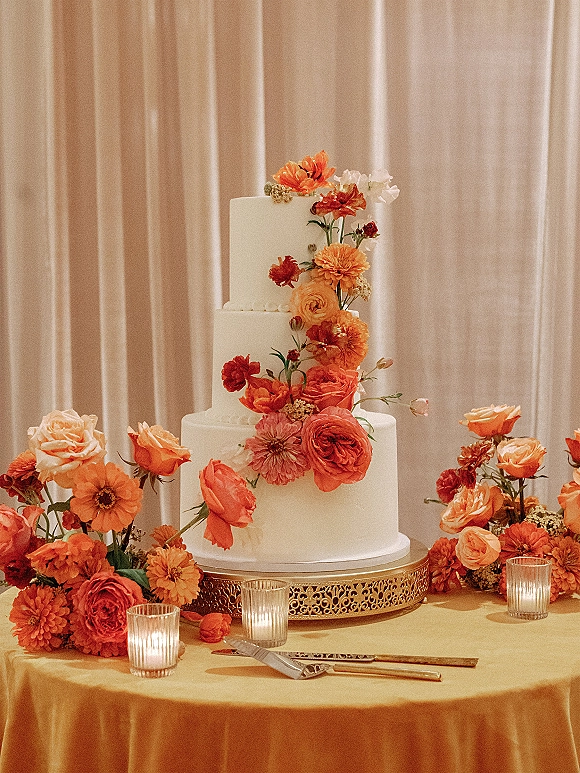 Wedding cake with orange roses and coral blooms on a gold stand, surrounded by votive candles on a yellow tablecloth before draped curtains