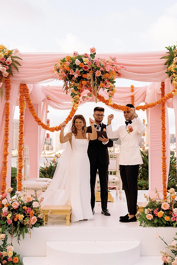 Wedding ceremony moment as bride and groom raise hands beside officiant under pink draped floral arch, with waterfront city skyline behind