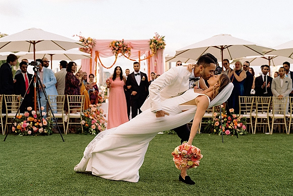 Wedding kiss portrait of groom dipping bride in veil as she holds bouquet, with floral arch, gold chairs, guests and umbrellas on lawn