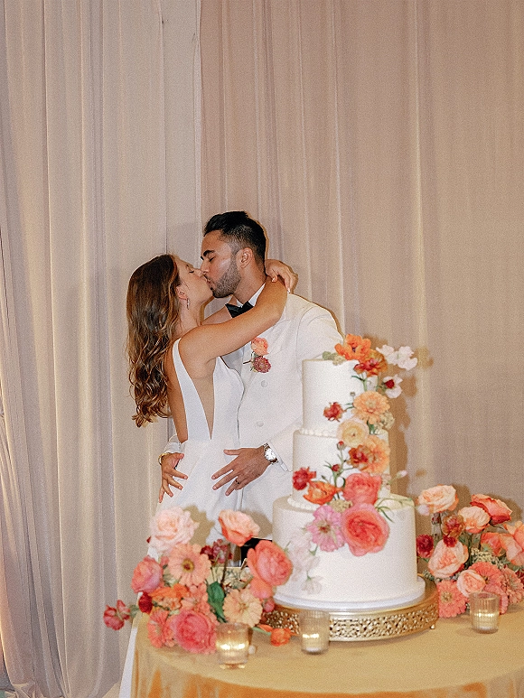 Wedding cake cutting as bride in backless dress and groom in tuxedo kiss behind three-tier cake with sugar flowers and draped backdrop