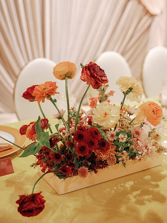 Wedding centerpiece with colorful blooms on a wooden tray, set among plates, napkins and place cards on a long table with draped backdrop