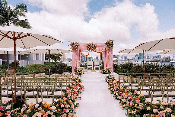 Ceremony setup with floral aisle runner and pink draped arch on a waterfront lawn, gold chiavari chairs under umbrellas, skyline beyond