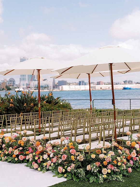 Ceremony seating setup with outdoor wedding chairs in gold chiavari rows, white cushions, floral aisle borders, and umbrellas by a waterfront skyline