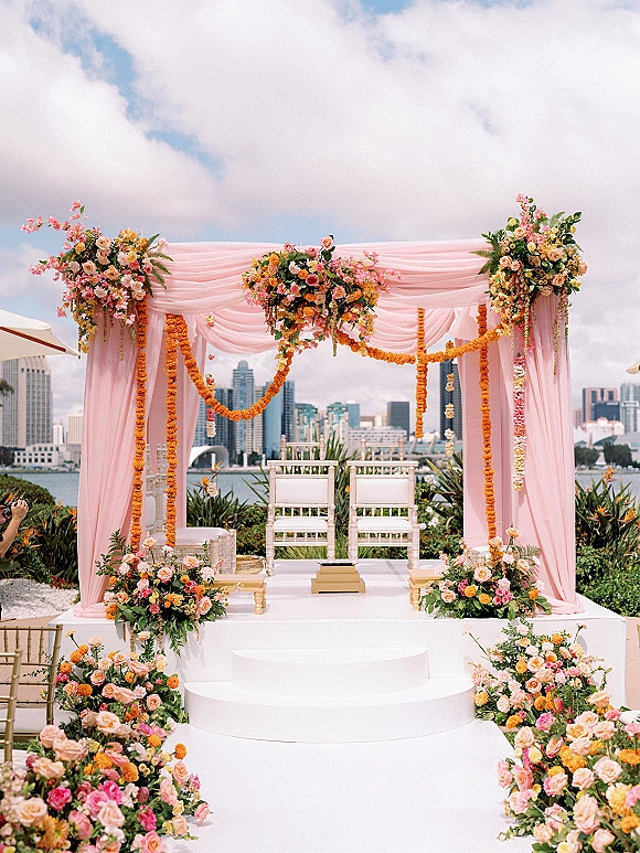 Wedding ceremony setup with pink draped mandap, pastel floral arch and marigold garlands on a white stage by the waterfront skyline