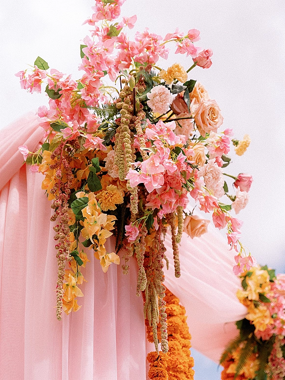 Wedding floral arrangement on a ceremony arch with pink and peach flowers, peach roses, hanging amaranthus, and draped fabric against a white sky