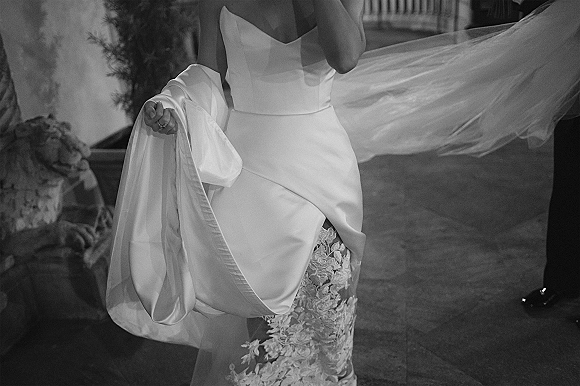 Wedding dress with a strapless gown and lace appliqué skirt, veil trailing beside a wedding ring on an indoor floor near a potted plant