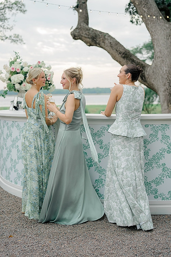Wedding guest dresses in sage green and pastel floral prints, long gowns with bow shoulder accents at a waterfront cocktail bar under string lights