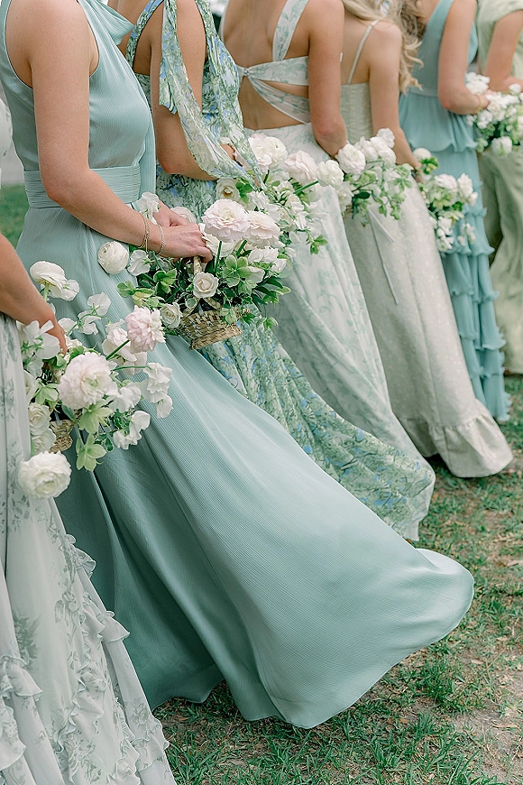Bridesmaid dresses in sage green with basket bouquets of white flowers and greenery, ribbon tie straps, standing on a grass lawn outdoors