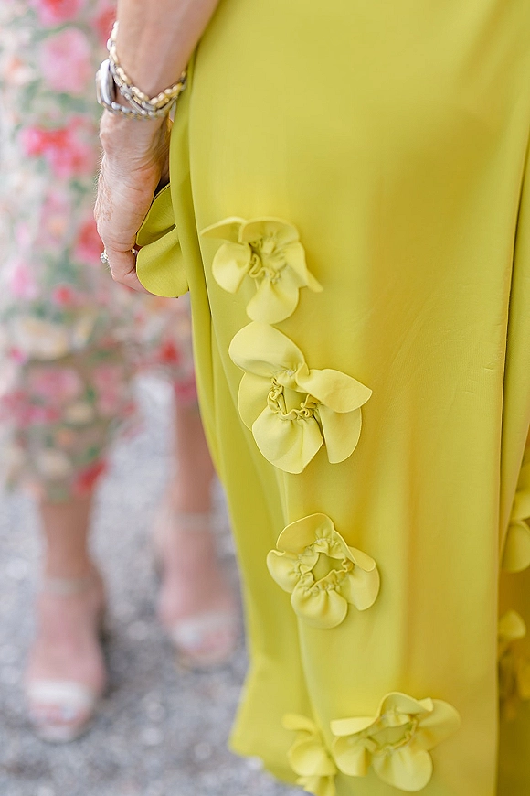 Bridesmaid dress detail on a chartreuse bridesmaid dress, showing 3D ruffle flower appliqués, bracelet stack, and ring near heels on gravel