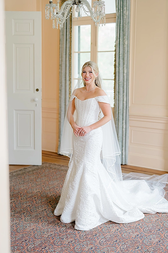 Bridal portrait of a bride in an off the shoulder wedding dress with cathedral veil, standing by tall windows under a chandelier indoors