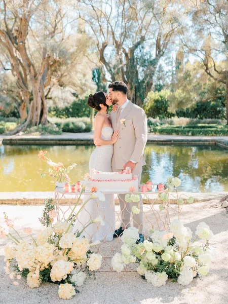 Wedding kiss portrait of bride and groom kissing beside a floral wedding cake table, with hydrangeas and calla lilies by a garden pond