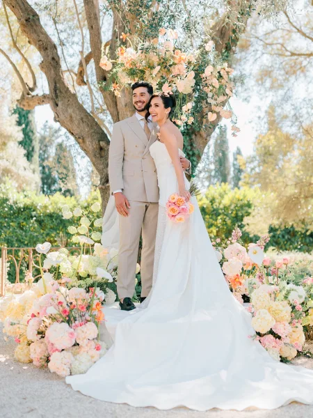 Couple portrait of bride and groom smiling under a floral arch, her strapless dress with long train and pastel bouquet on a sunlit garden path