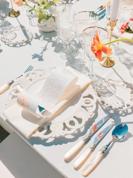 Reception tablescape wedding place setting with white cutwork charger, menu on napkin, gold candlesticks, and orange flower stem in sunlit shadows