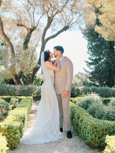 Couple portrait of bride and groom embracing, bride in long veil touching his face, on a sunlit garden path with trees and hedges