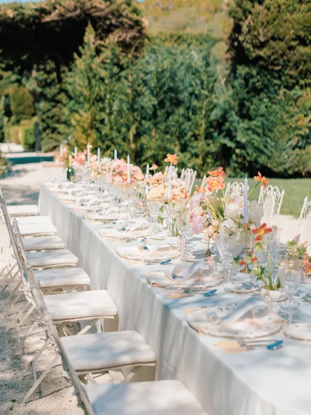 Reception tablescape on a long outdoor wedding reception table with pastel florals, taper candles, patterned plates, and menus in sunlit garden greenery