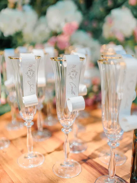 Wedding place cards displayed on champagne flutes with calligraphy name tags and gold-rim glassware on a wooden table before a floral wall backdrop