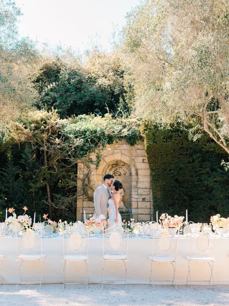 Wedding couple portrait of bride and groom kissing beside a long banquet table with pink and peach florals, candles, and a stone fountain courtyard
